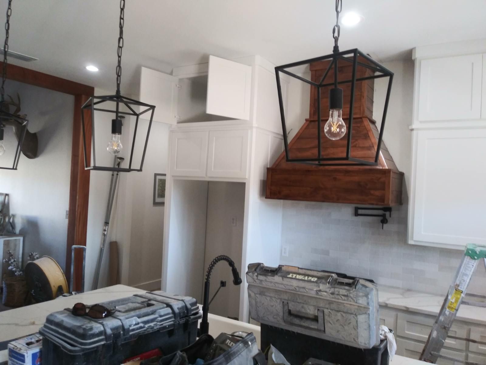 Kitchen remodel with hanging lights, wooden hood, white cabinets, tools on counter.