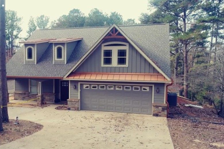 Gray house with a copper-colored roof over the garage and two dormers. Stone accents, trees in the background.