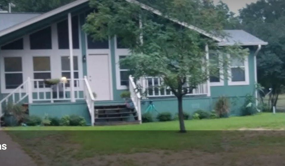 Green house with white trim and a porch; small tree in front.