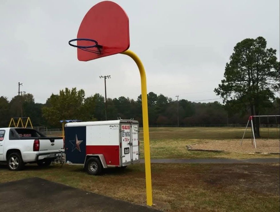 Basketball hoop with a red backboard, yellow pole, and a trailer with a Texas flag on a cloudy day in a park.