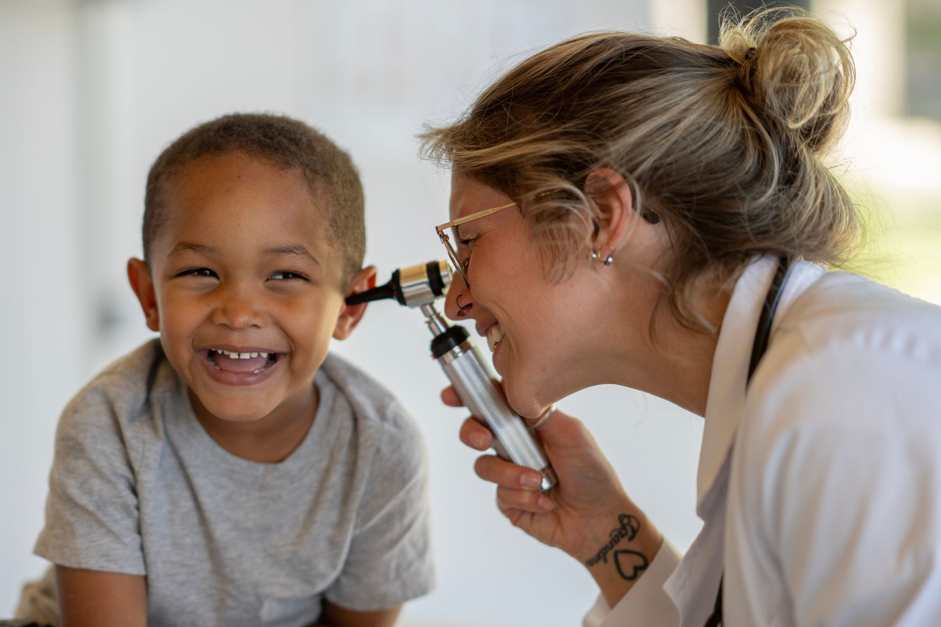 Doctor examining a smiling child's ear with an otoscope. White lab coat, light skin.
