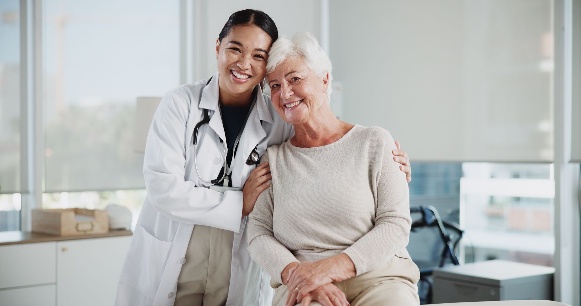 A young doctor with stethoscope, smiling, hugs an older patient in a brightly lit office.