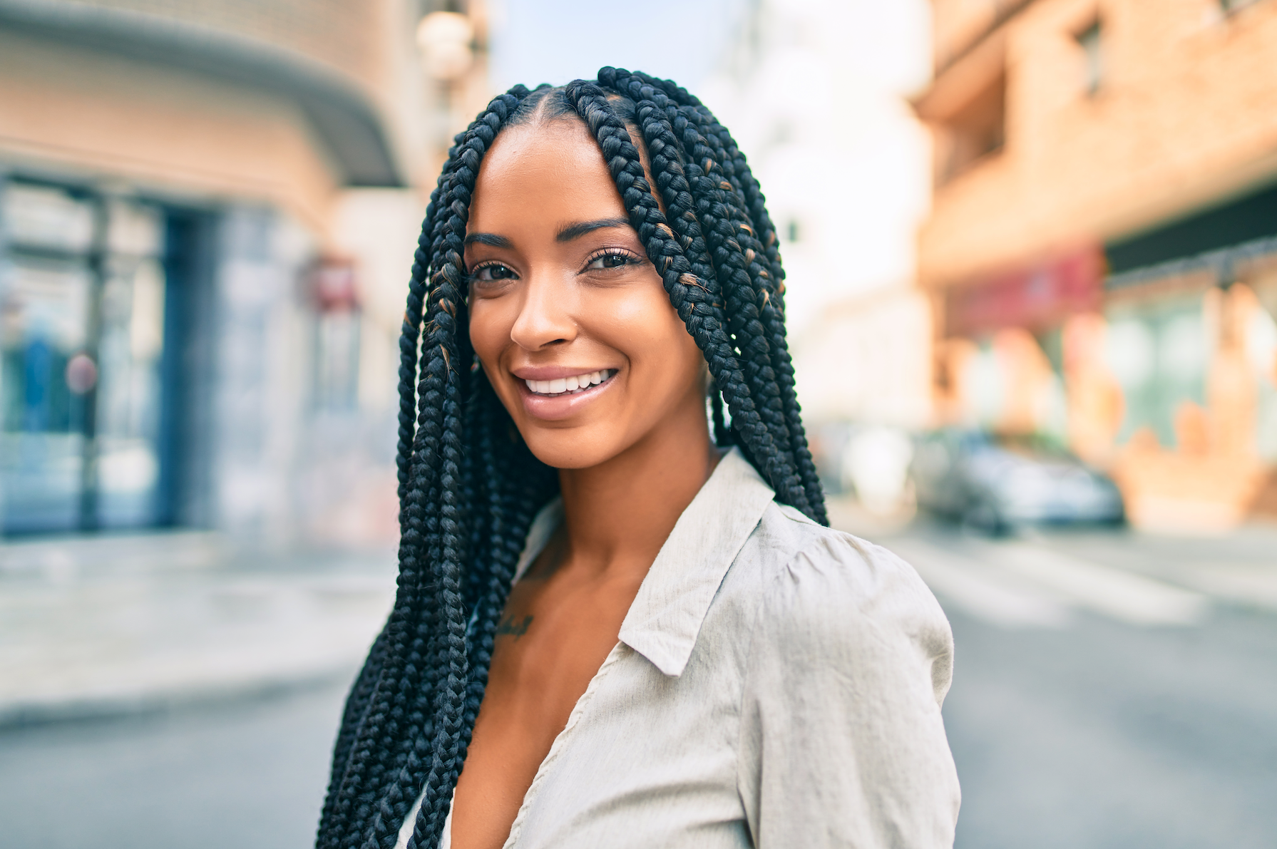A woman with braids is smiling and looking at the camera while standing on a city street.