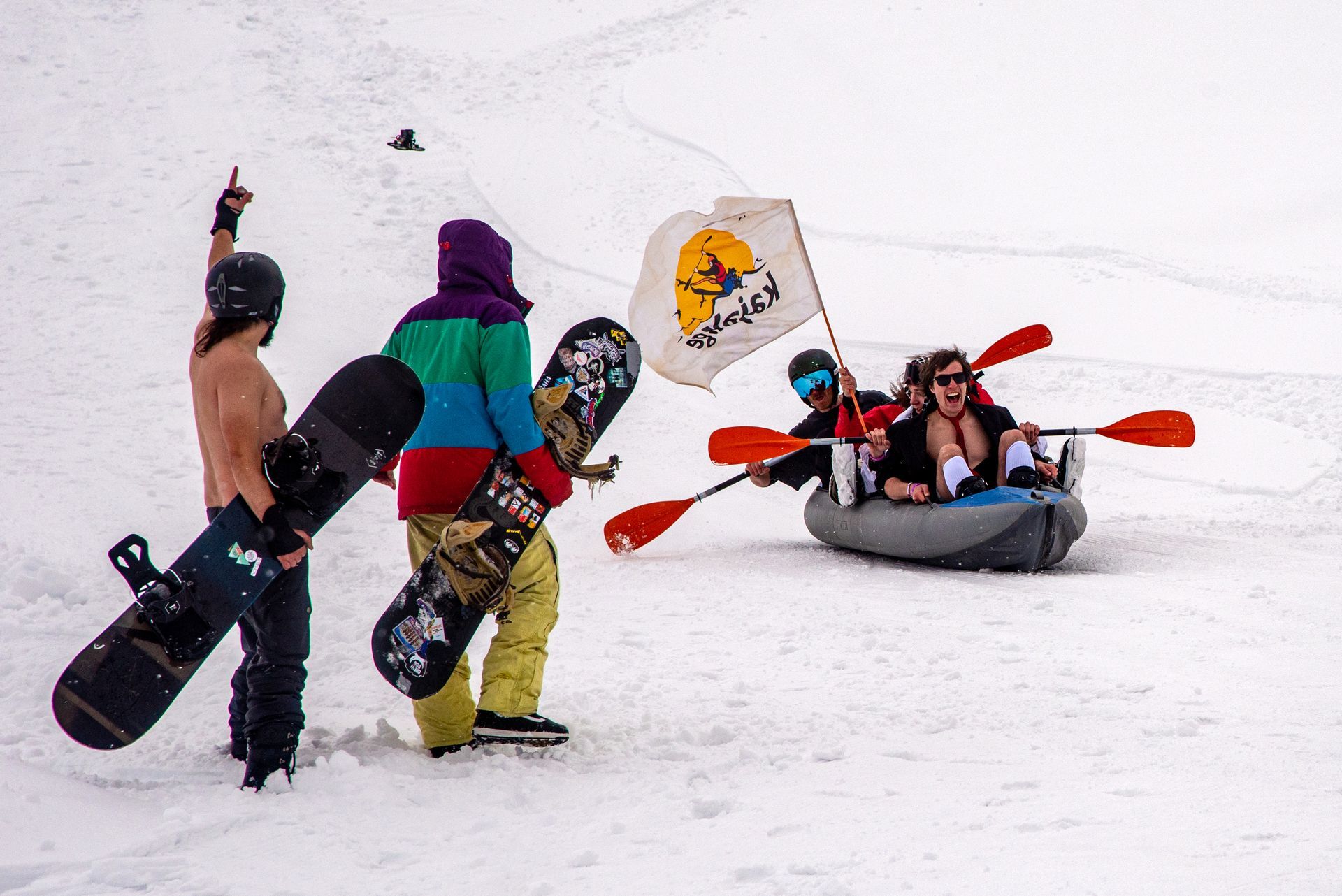 People with snowboards and paddles interact near a raft on a snowy slope.