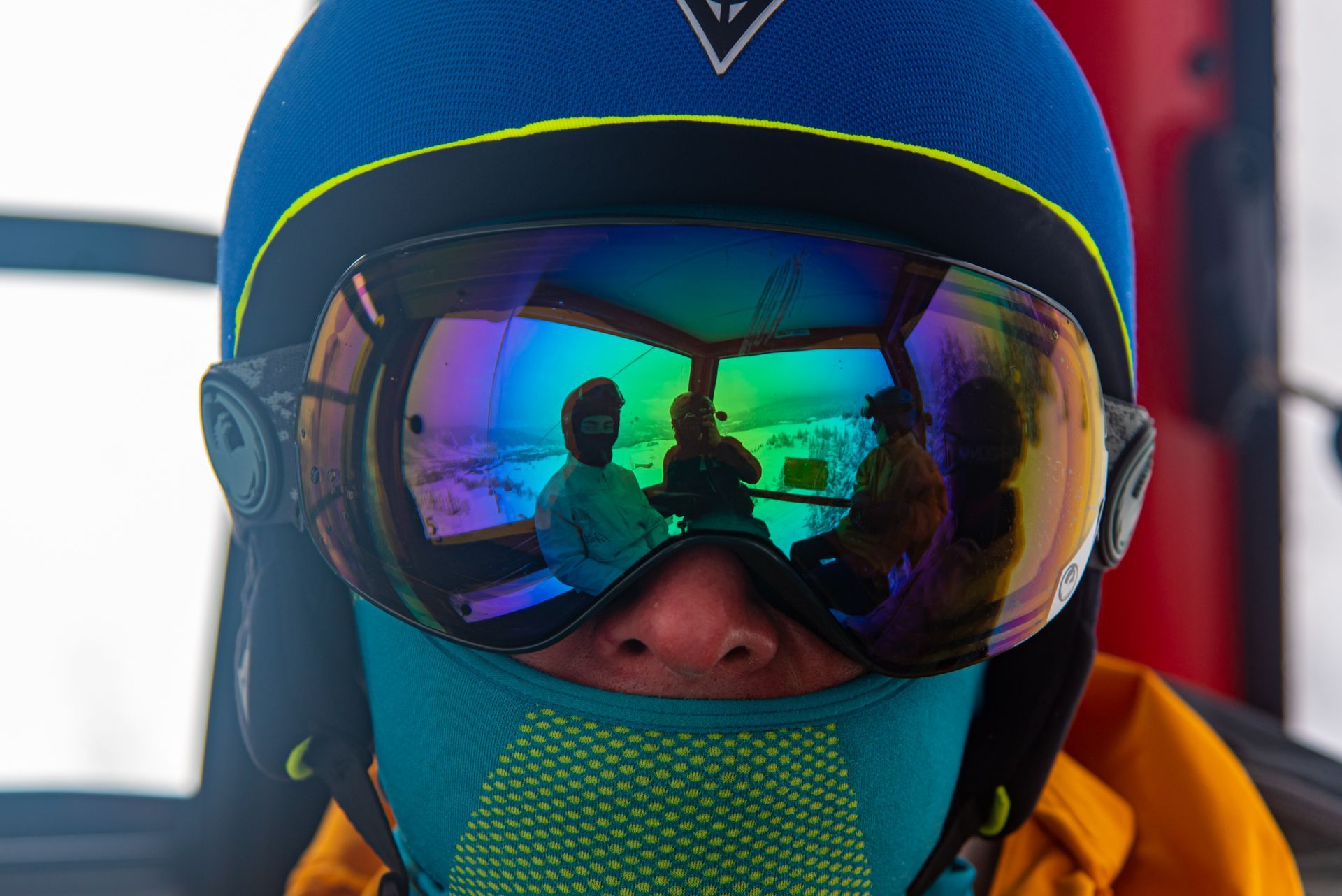 Person in blue ski helmet and goggles, reflection of three people inside a vehicle.