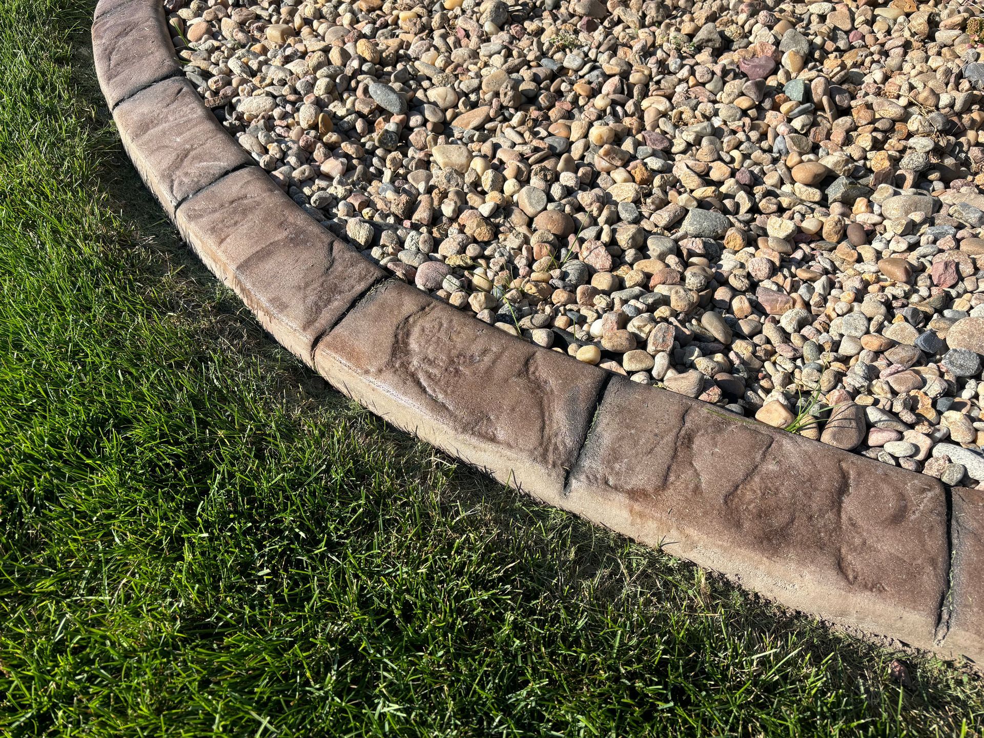 Curved stone-like border separates green grass from a bed of small brown and grey gravel.
