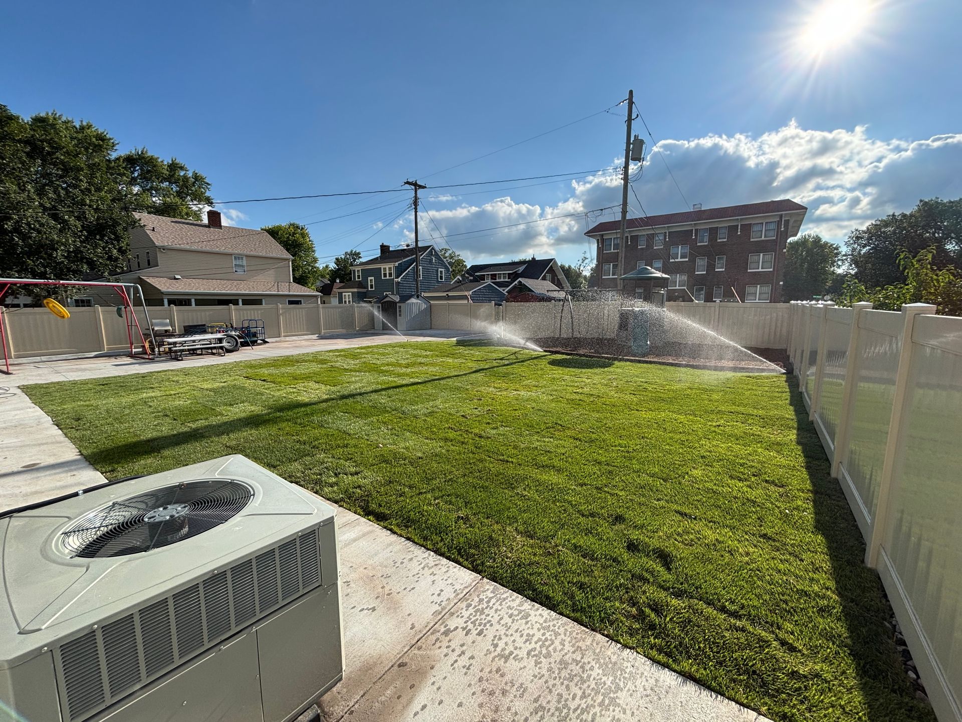 Sprinklers watering a newly planted lawn enclosed by a white fence on a sunny day.