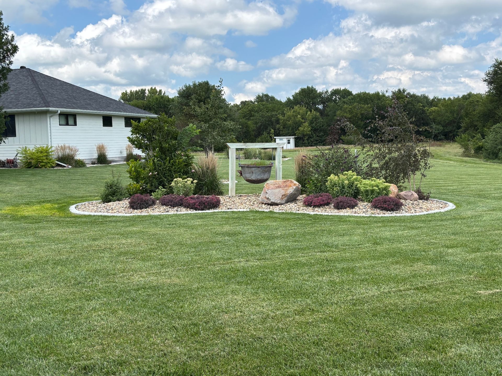 Lawn with a flower bed, large rocks, and a swing set under a cloudy sky near a house and trees.