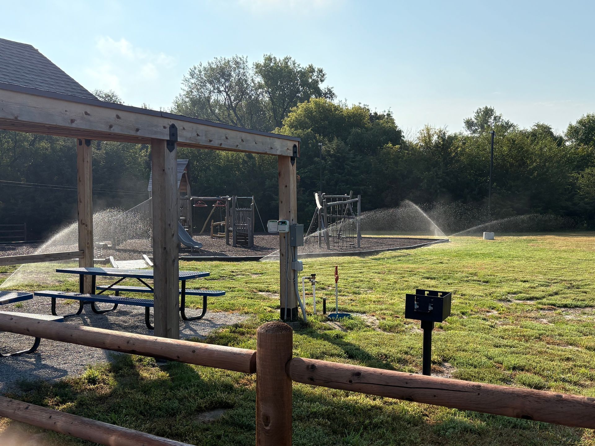 Picnic area with sprinklers running, playground in the background, and a wooded area behind it.