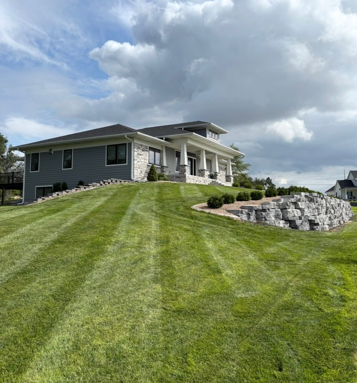 Modern house on a grassy hill with stone retaining wall. Cloudy sky. Freshly cut lawn.