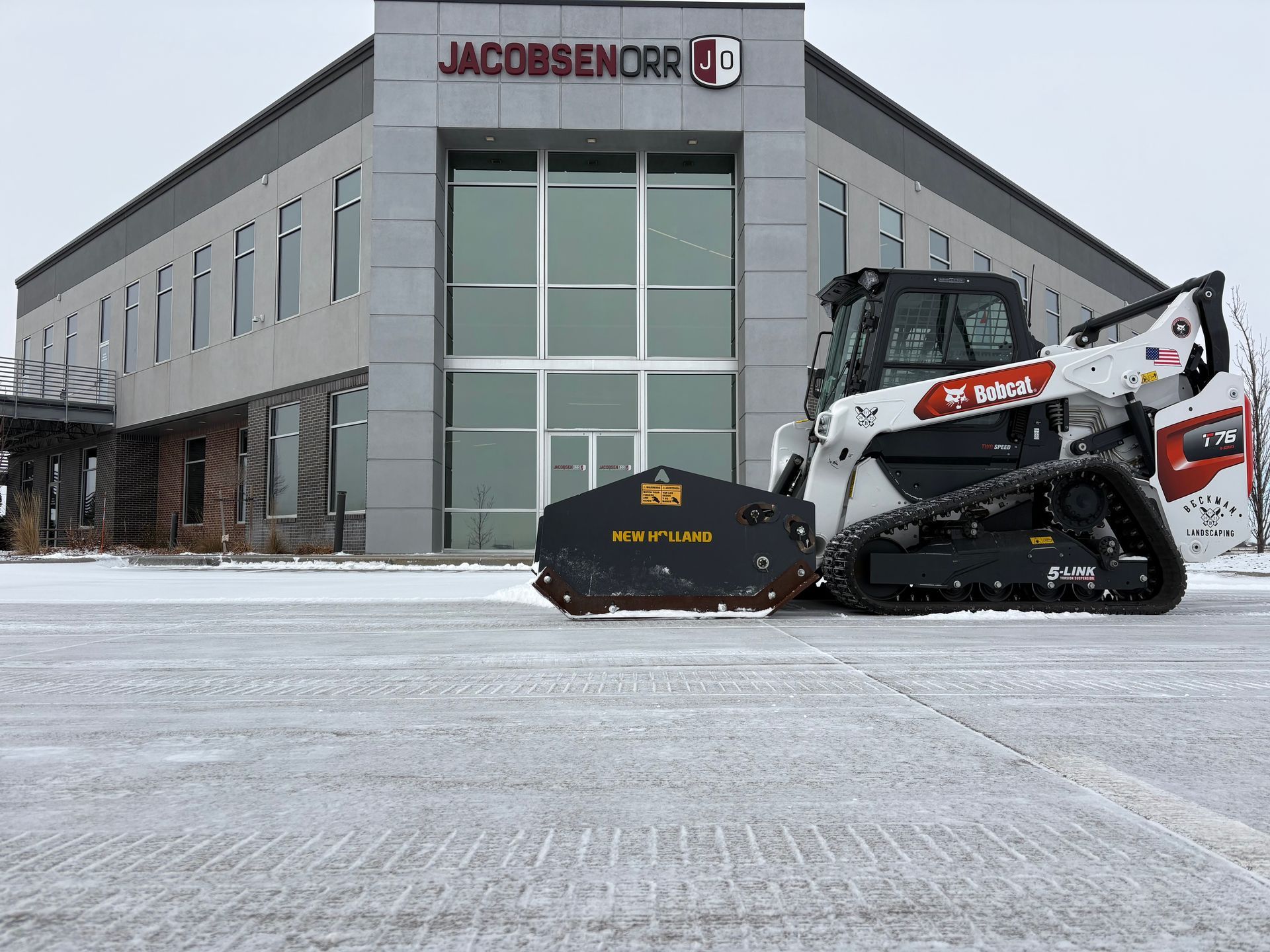 A Bobcat skid-steer with a brush cutter in front of a Jacobsen Orr building on a snowy day.