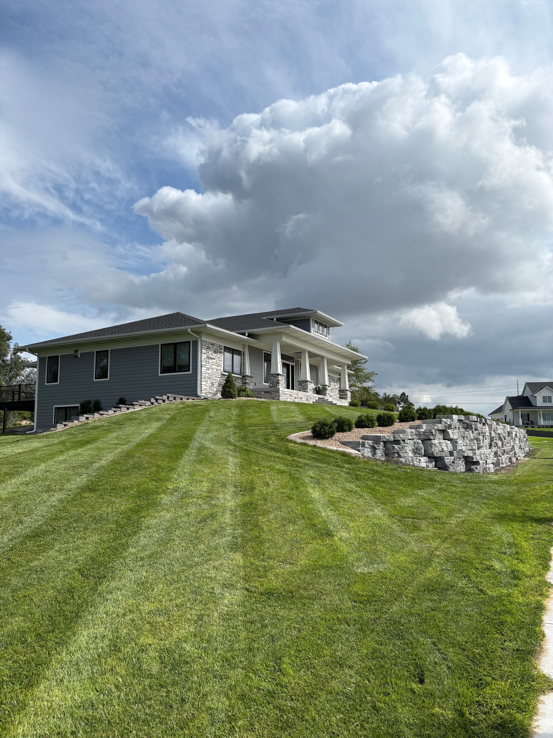 House atop a hill with mowed grass and cloudy sky. Stone retaining wall on right.
