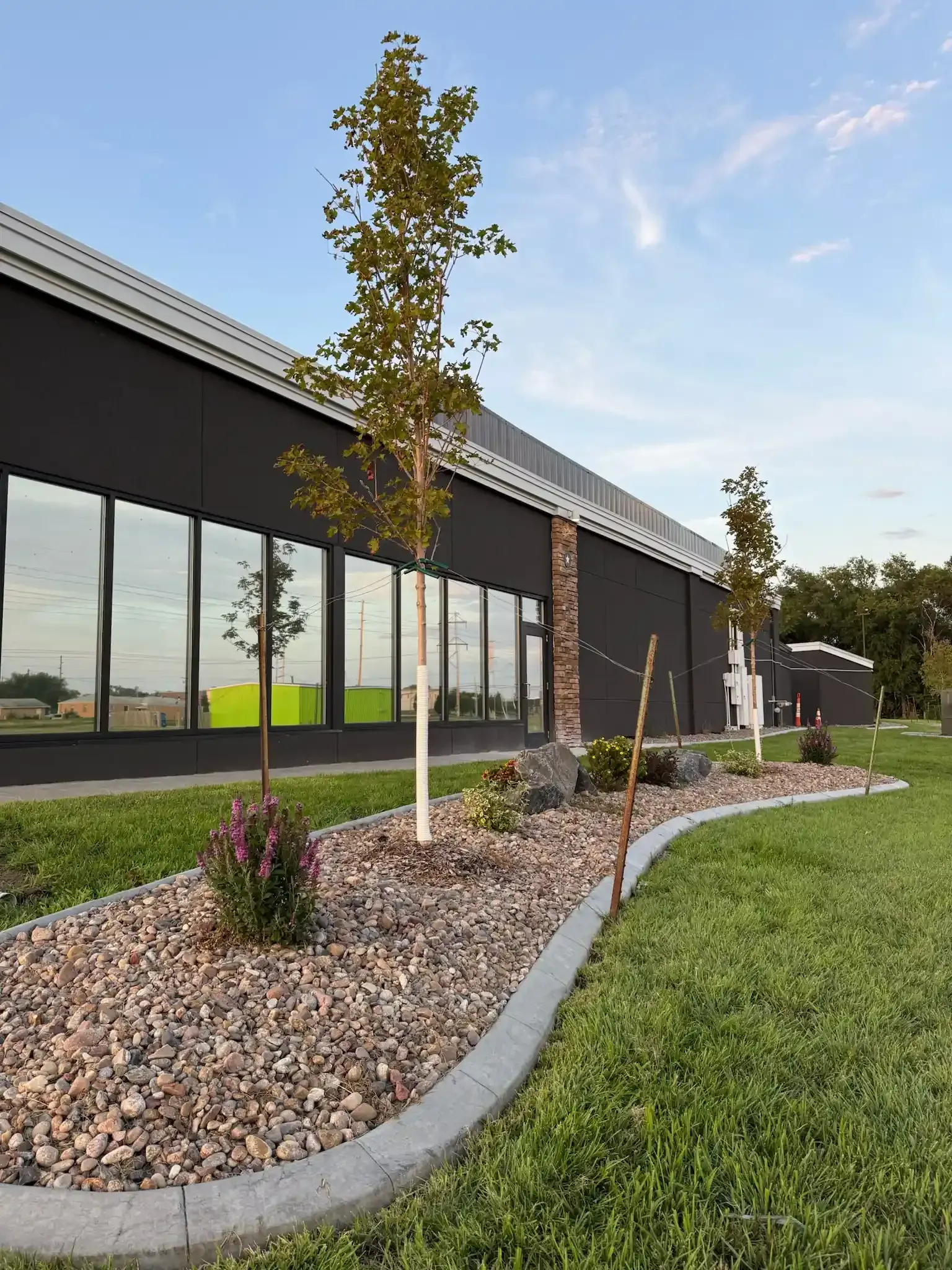 Modern building with dark facade, large windows, and landscaped bed with trees and rocks.