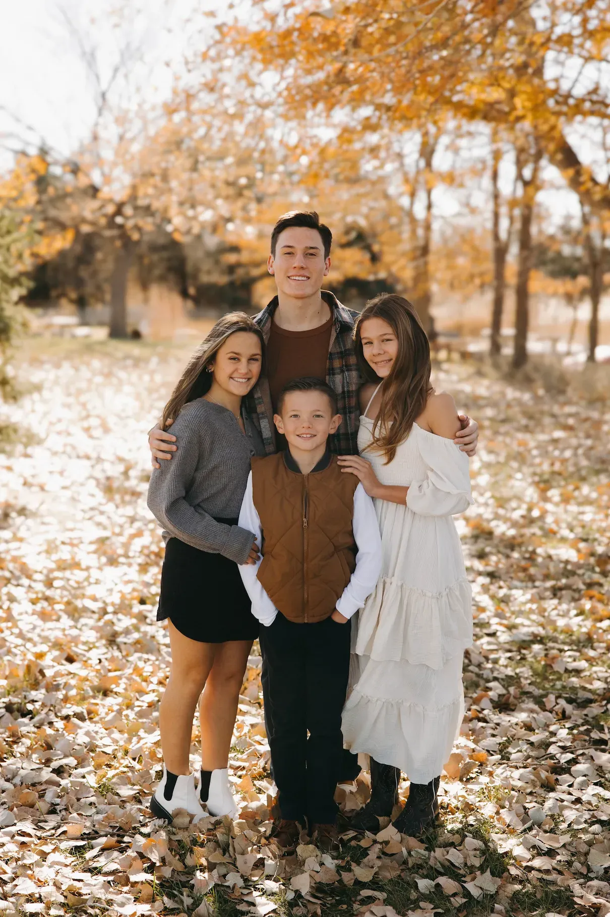 Family of five smiles while standing outdoors in autumn leaves.
