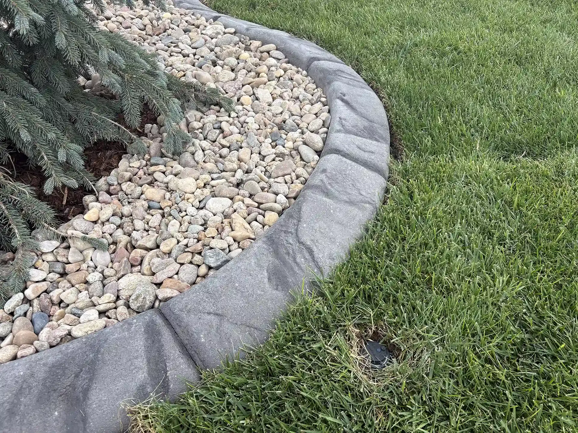 Gray stone curbing separates a flowerbed with rocks from green grass.