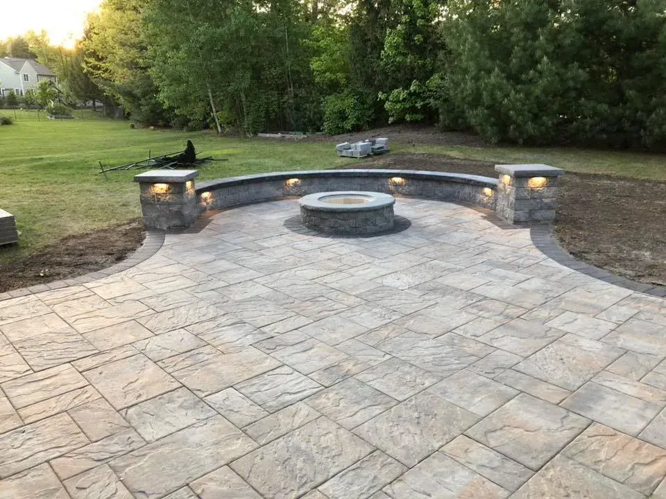 Stone patio with built-in fire pit, surrounded by a curved wall with lighting. Lawn and trees in background.