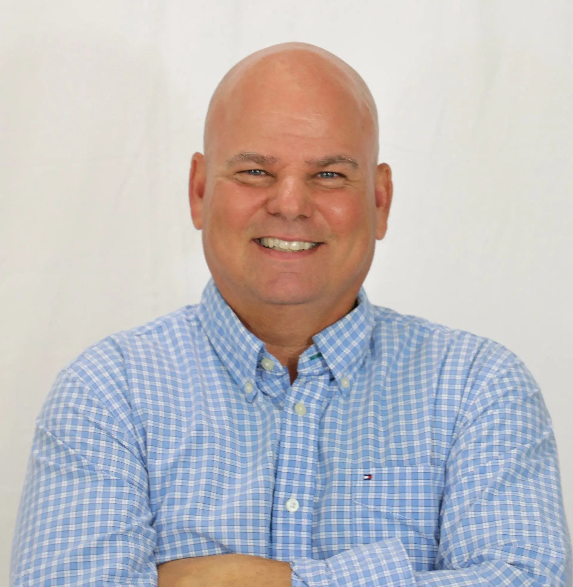 A bald man in a blue plaid shirt is smiling with his arms crossed.
