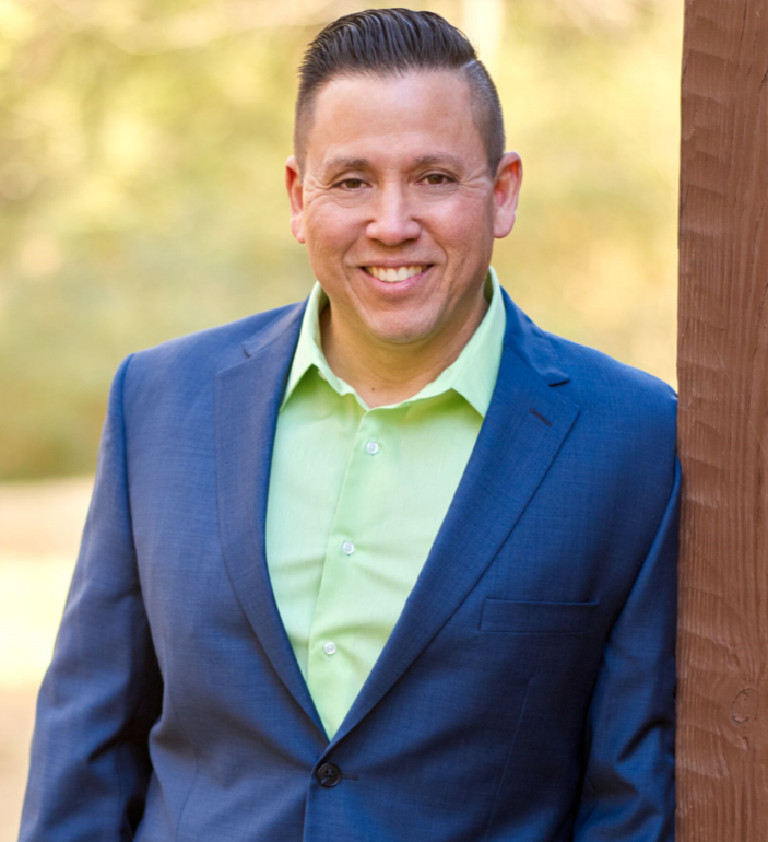 A man in a blue suit and green shirt is leaning against a wooden post
