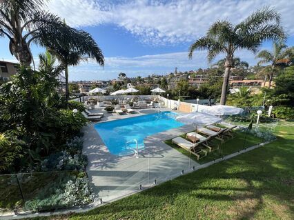 Resort pool with lounge chairs, palm trees, and ocean view under a bright blue sky