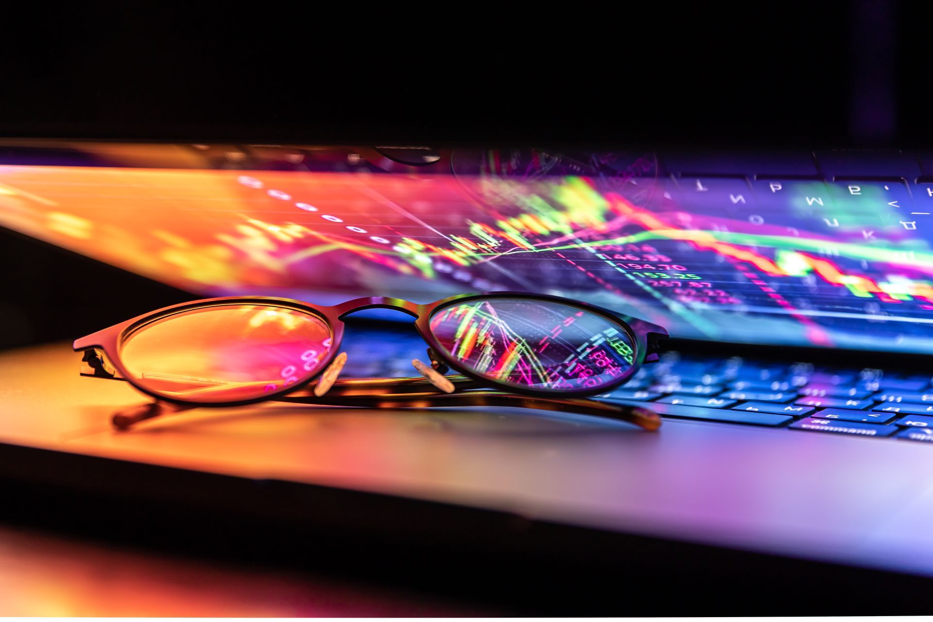 Glasses on a laptop with a colorful stock market graph displayed on the screen.