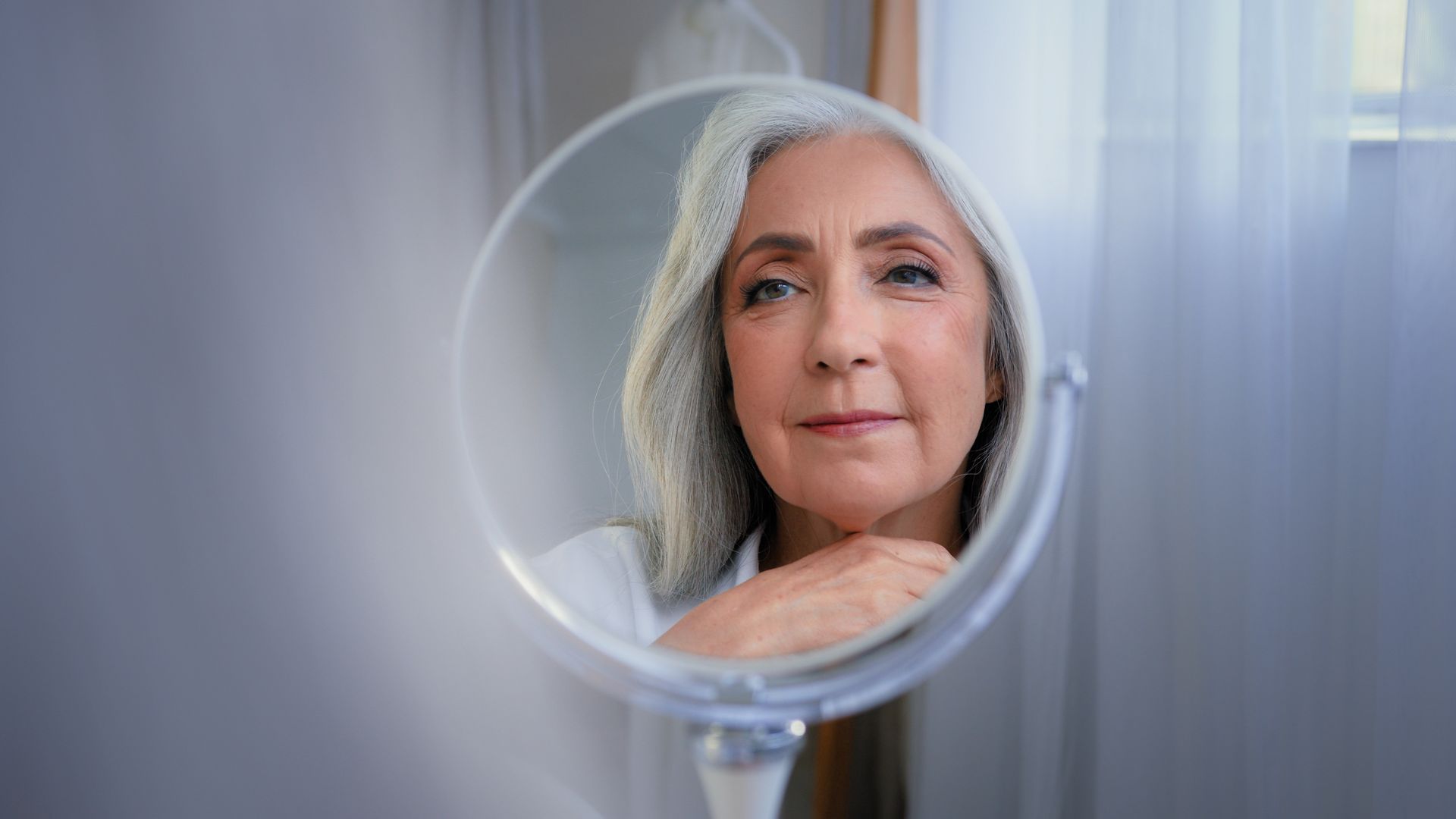 Mature woman with gray hair looking at herself in a round mirror, touching her neck. Neutral background, soft lighting.