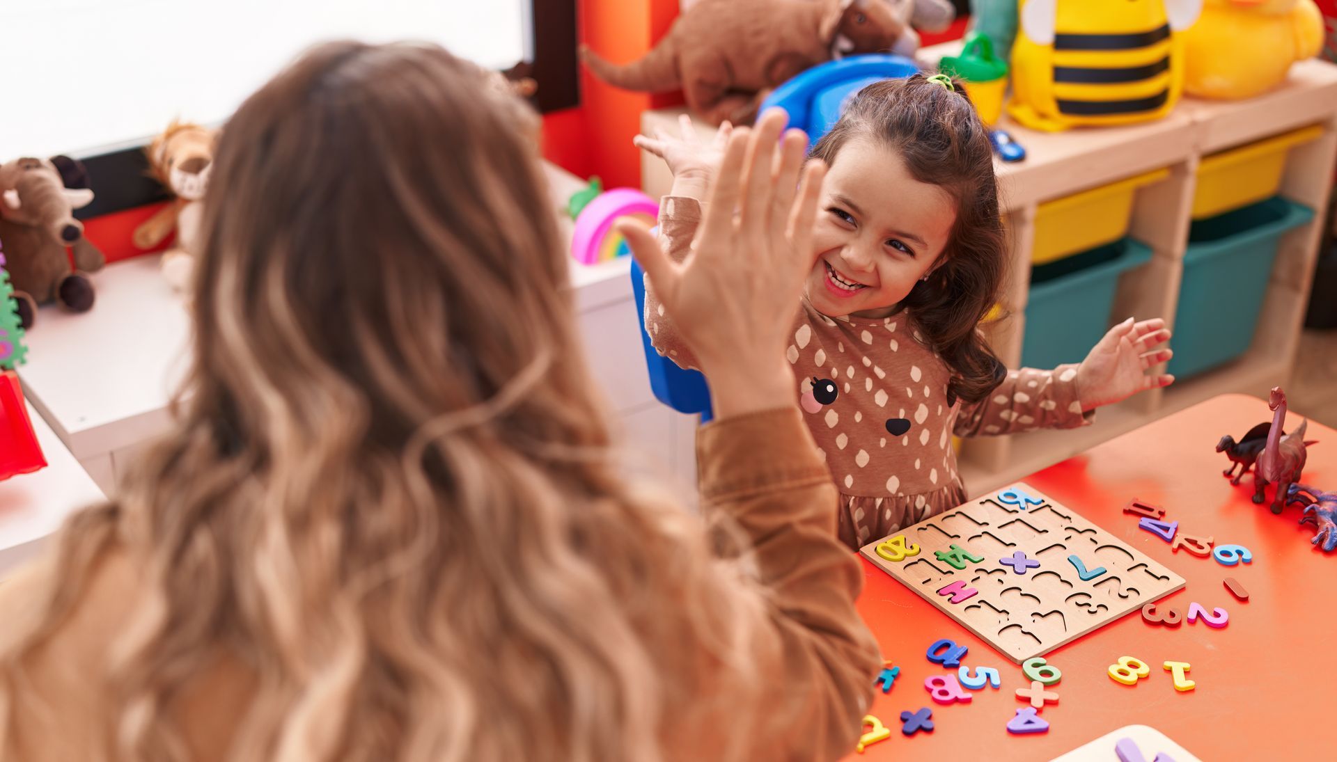 Lehrerin und Kind geben sich an einem Klassenzimmertisch mit bunten Buchstaben und Spielzeug ein High-Five; das Kind lächelt.
