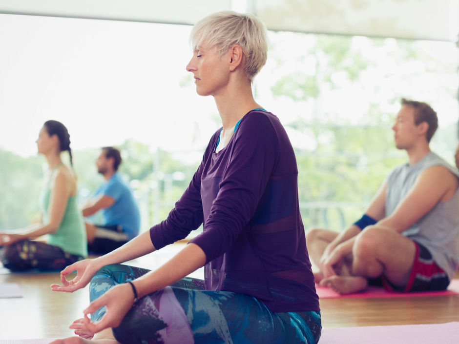 Menschen meditieren auf Yogamatten in einem hellen Studio; im Vordergrund ist eine Frau in einem lila Oberteil zu sehen.