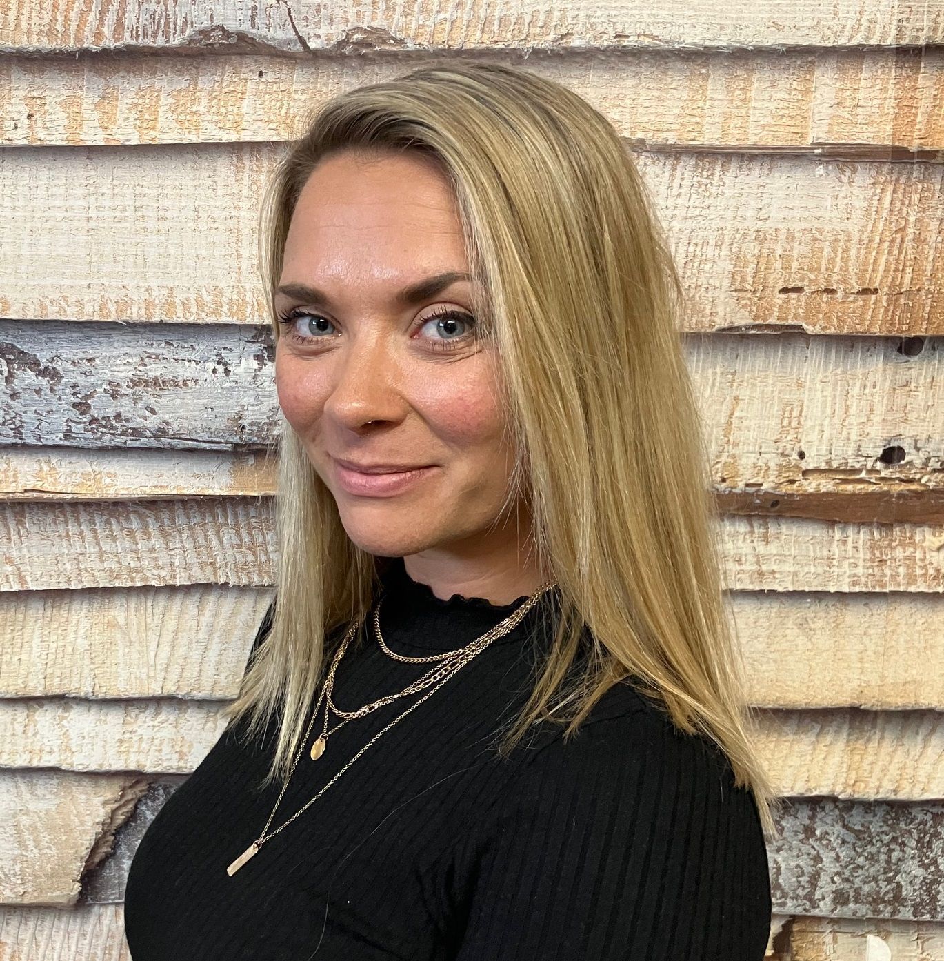 Blonde Frau in schwarzem Oberteil und mehreren Halsketten steht vor einer strukturierten Holzwand und lächelt leicht.