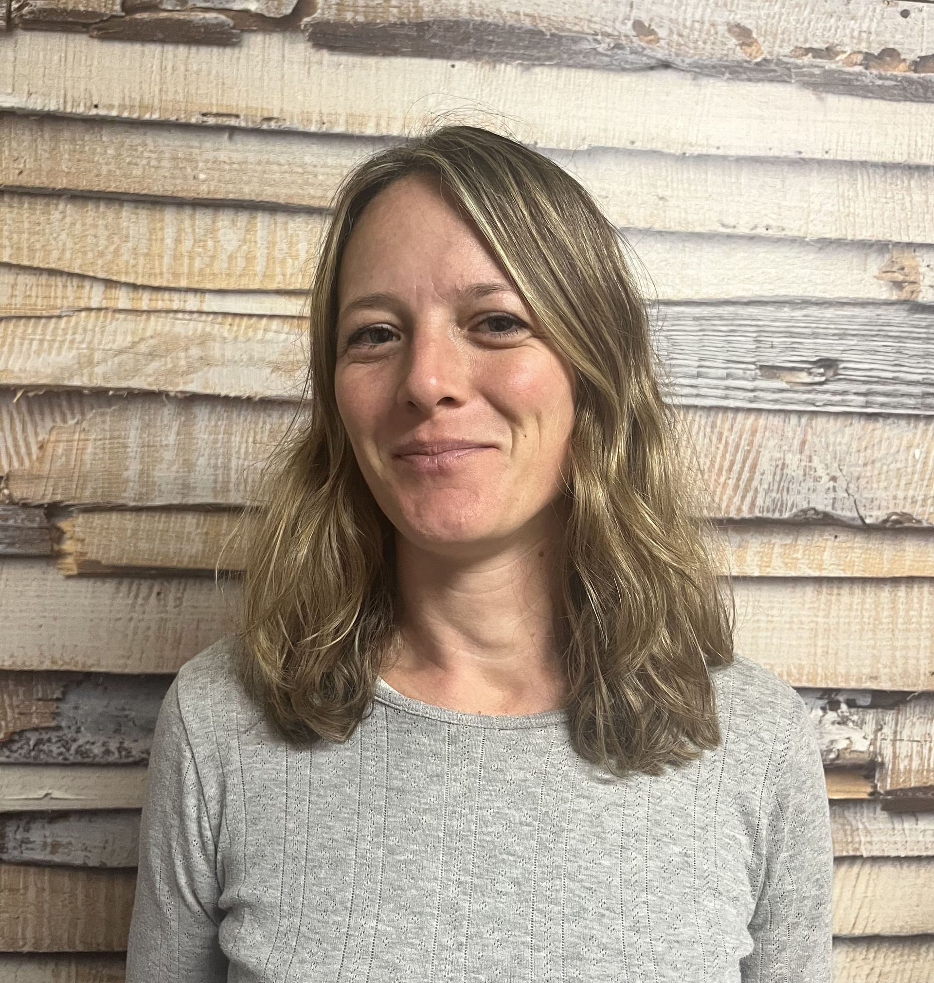 Lächelnde Person mit schulterlangem Haar in einem grauen Pullover vor hellem Holzplankenhintergrund