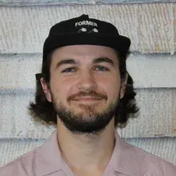 Eine lächelnde Person mit schwarzer Kappe und hellrosa Hemd vor einer strukturierten Wand