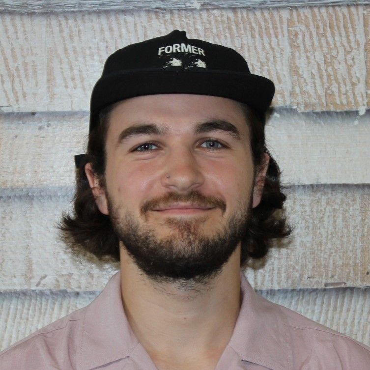 Porträt einer lächelnden Person mit schwarzer Kappe und rosa Hemd vor einer hellen Holzwand