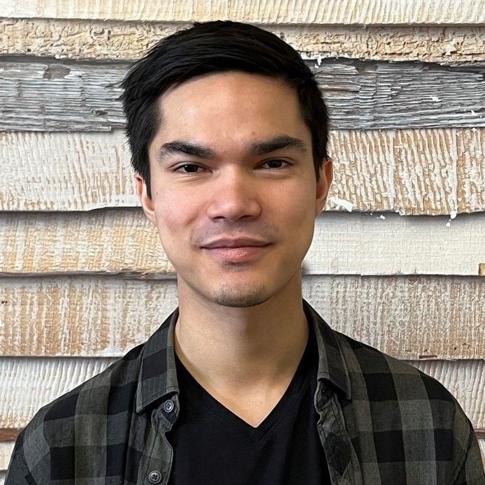 Lächelnde Person in schwarzem Hemd und karierter Jacke vor einer strukturierten Holzwand