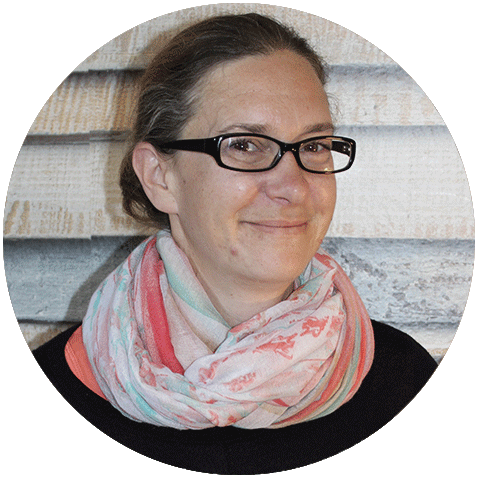 Porträt einer Person mit Brille und rosa gemustertem Schal, die vor einer strukturierten Wand leicht lächelt.