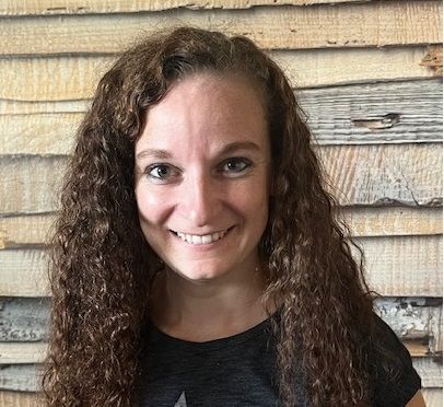 Lächelnde Frau mit lockigem braunem Haar in einem schwarzen Hemd vor einer strukturierten Holzwand