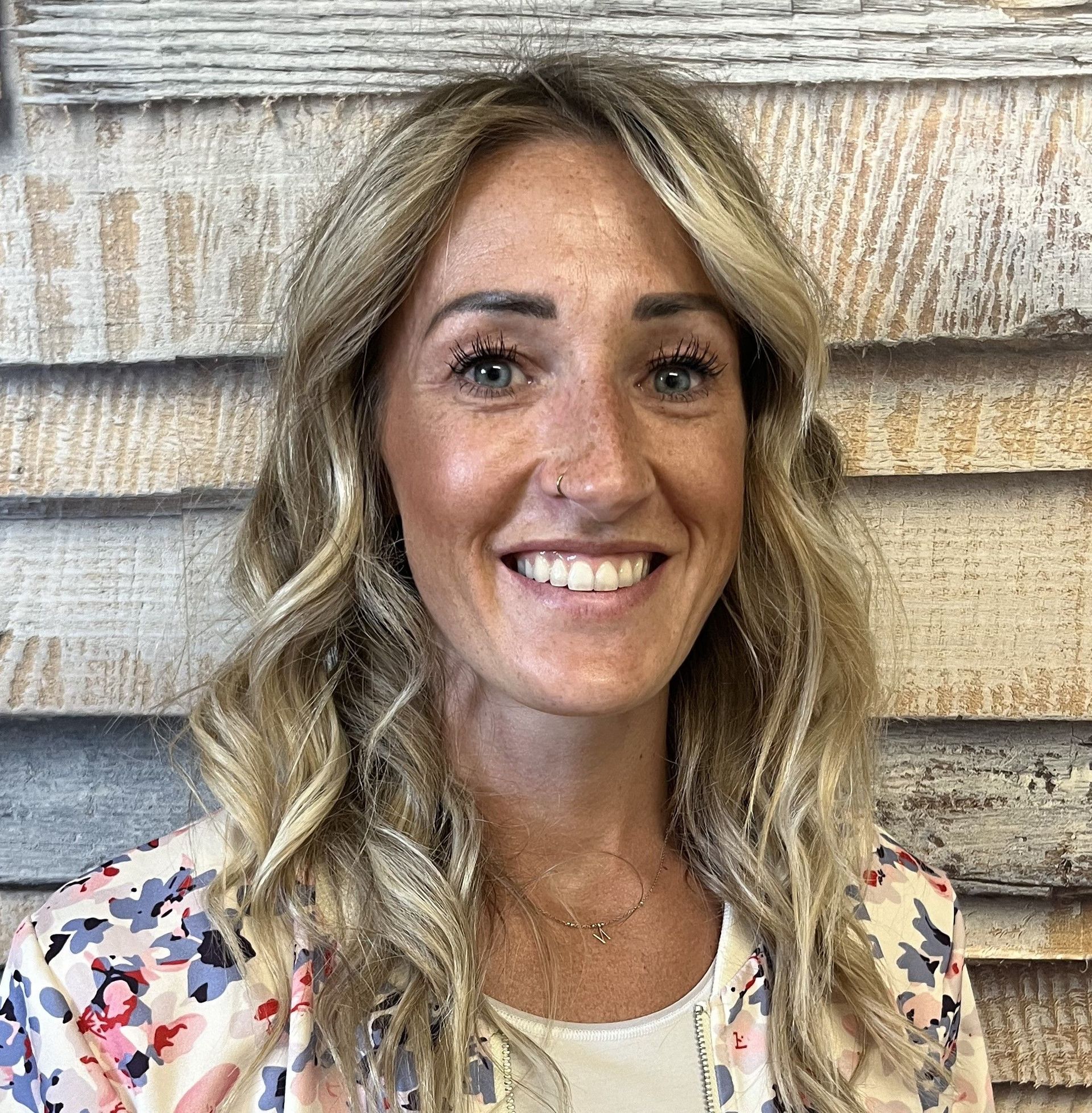 Lächelnde Person mit welligem blonden Haar und einem geblümten Oberteil vor einer verwitterten Holzwand