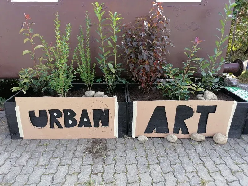 Pflanzkästen mit den Aufschriften „URBAN“ und „ART“, bepflanzt mit grünen und violetten Pflanzen an einer Wand.