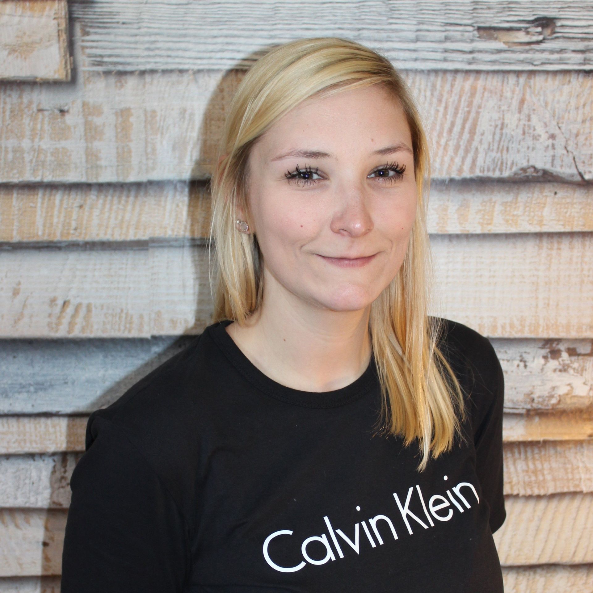 Frau in einem schwarzen Calvin Klein Hemd, lächelnd vor einer rustikalen Holzwand.