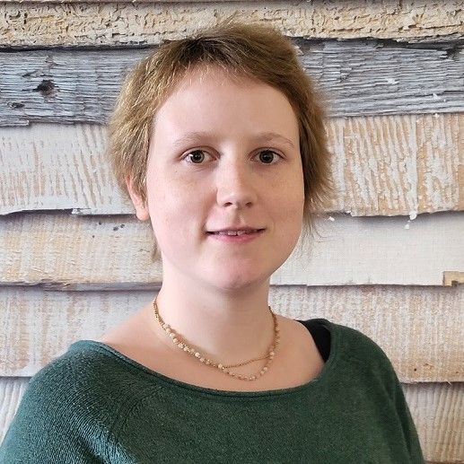 Porträt einer Person mit kurzen, hellen Haaren, die einen grünen Pullover trägt, vor einer Holzwand.