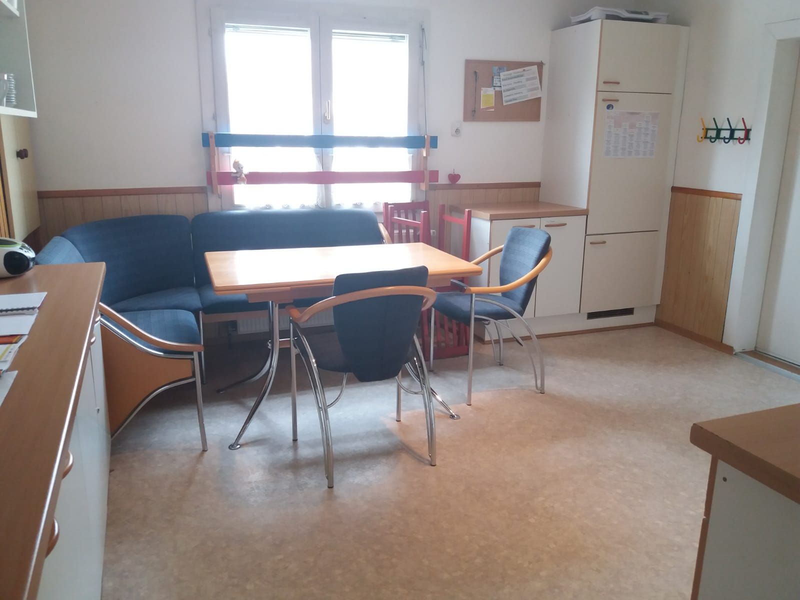 Kleiner Pausenraum im Büro mit einer blauen Couch, einem Tisch, Stühlen und einem Schrank am Fenster.