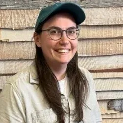 Eine lächelnde Person mit Brille und grüner Kappe steht an einer Schindelwand.