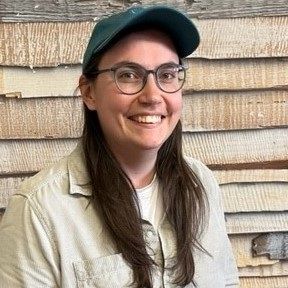 Eine lächelnde Person mit Brille und grüner Kappe steht vor einer rustikalen Schindelwand.