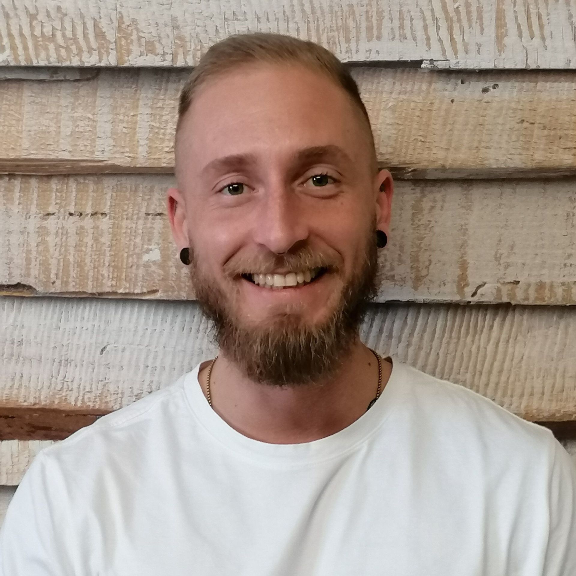 Lächelnder Mensch mit kurzen Haaren und Bart, der ein weißes Hemd trägt, vor einer strukturierten Holzwand.
