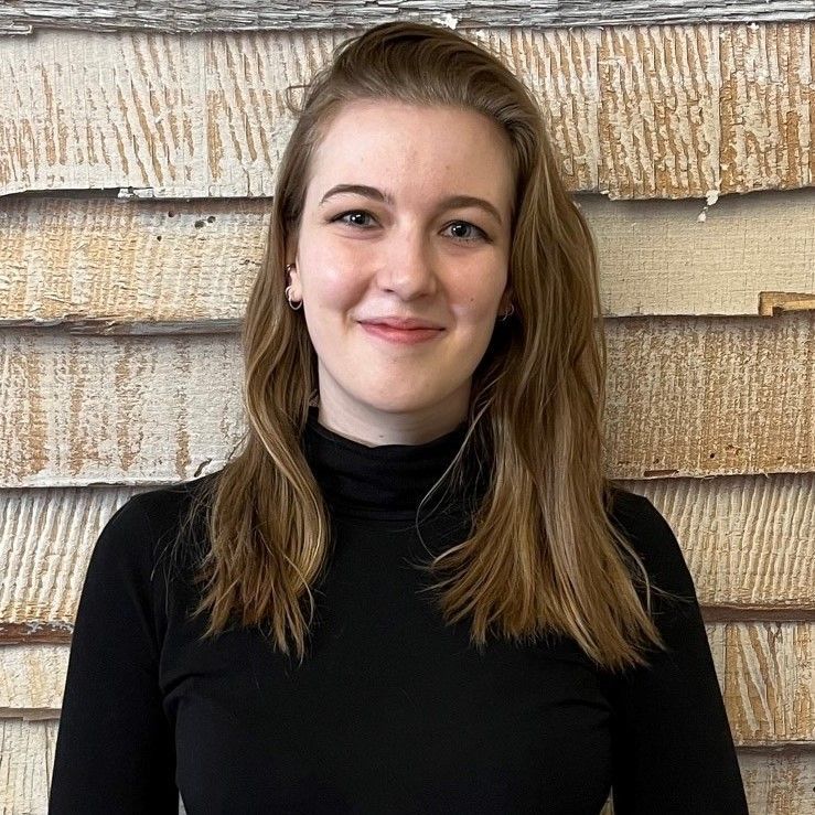 Porträt einer lächelnden Person in einem schwarzen Rollkragenpullover vor einer rustikalen Holzwand