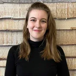 Eine lächelnde Person in einem schwarzen Oberteil vor einer strukturierten, hellbraunen Wand