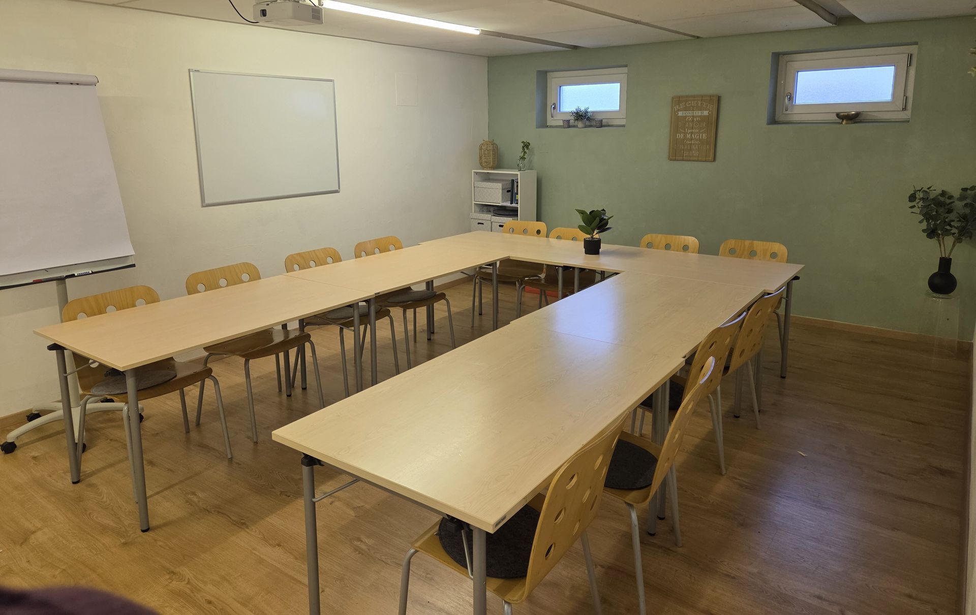 Leerer Konferenzraum mit U-förmigen Holztischen, Stühlen, Whiteboard und kleinen Fenstern.