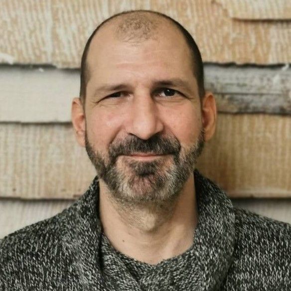 Lächelnder, kahlköpfiger Mann mit grau meliertem Bart vor einer strukturierten Holzwand