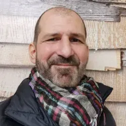 Ein lächelnder Mann mit kariertem Schal steht vor einer verwitterten Holzwand.