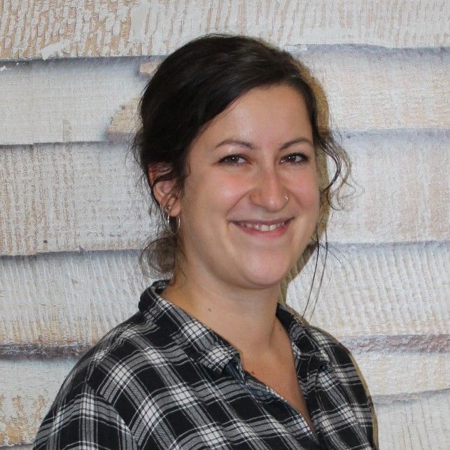 Lächelnde Person in einem schwarz-weiß karierten Hemd vor einer hellen Holzwand