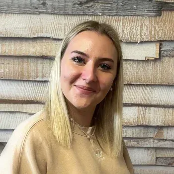 Lächelnde blonde Frau in einem beigen Oberteil vor einer strukturierten Holzwand.