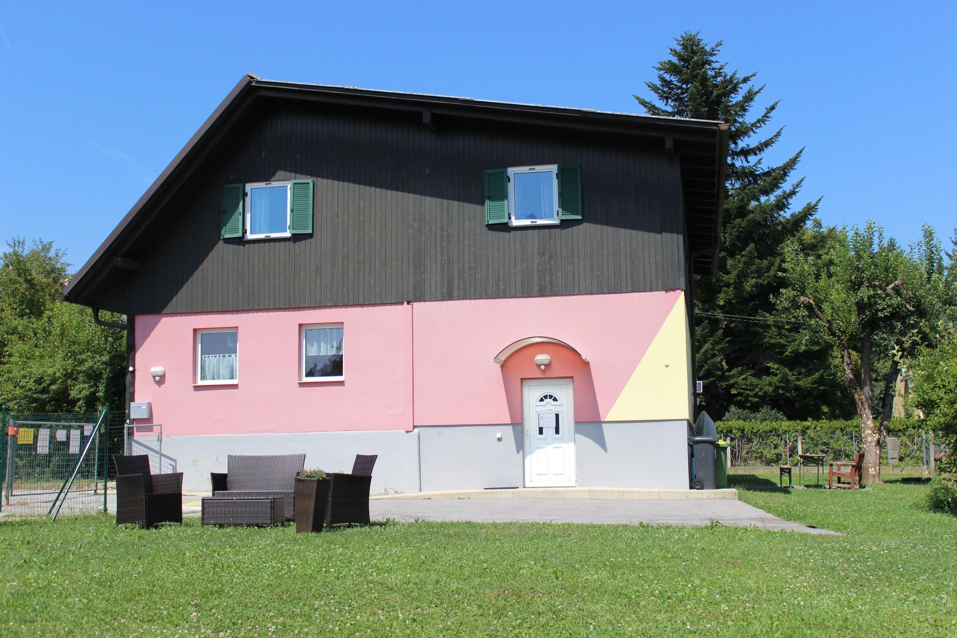 Ein rosa-graues Haus mit dunklem Dach und grünen Fensterläden auf einem sonnigen Rasen